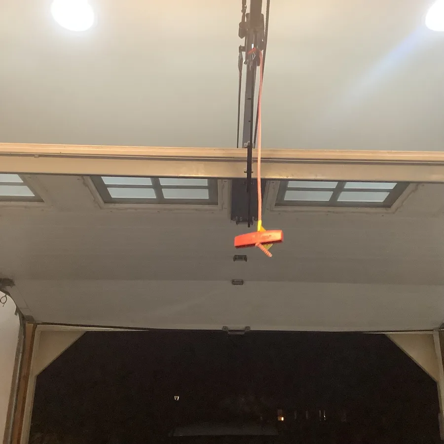Red emergency release handle hanging on its cord from the opener trolley at the header, above a garage door section with a row of small windows