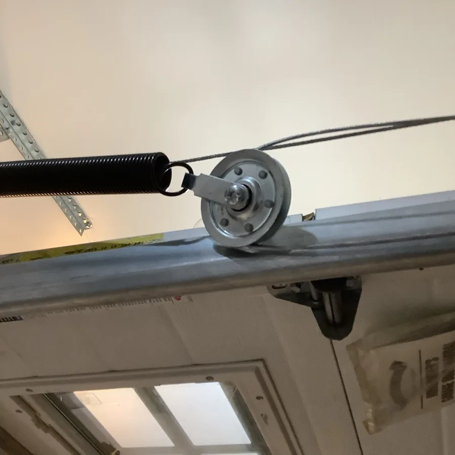 Extension spring hooked to a steel pulley sheave with lift cables running over it, sitting on the horizontal garage door track near the ceiling