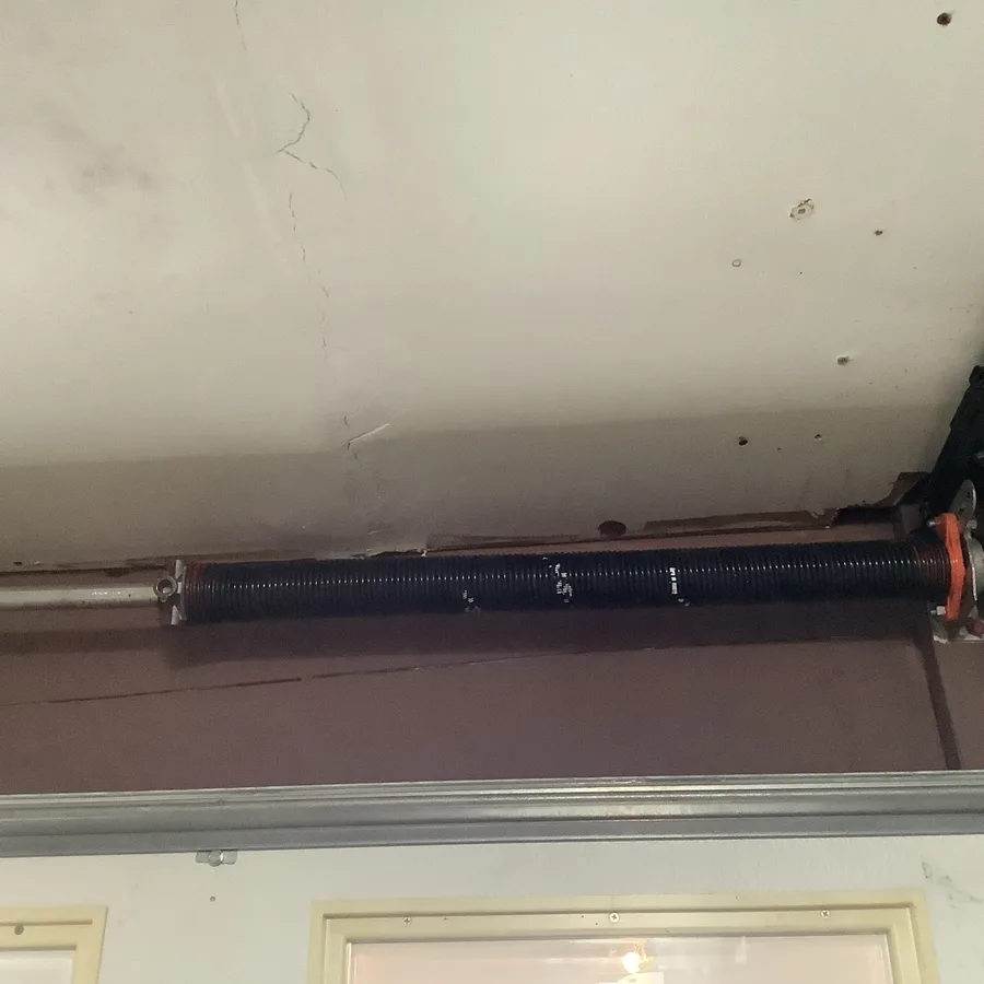 Torsion spring wound on its shaft running the width of the header above a garage door, mounted against a cracked painted ceiling