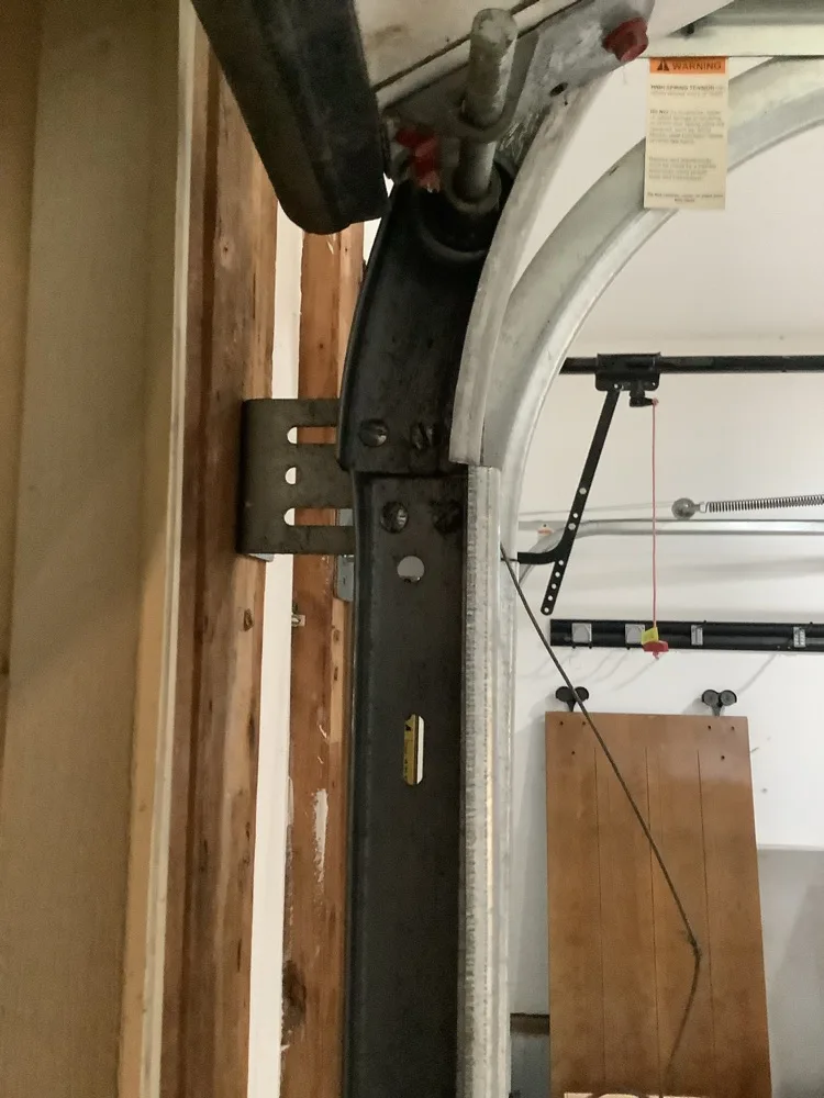 Vertical garage door track curving into the horizontal at the jamb, bolted to a timber frame through a slotted bracket, with a printed high spring tension warning label above it
