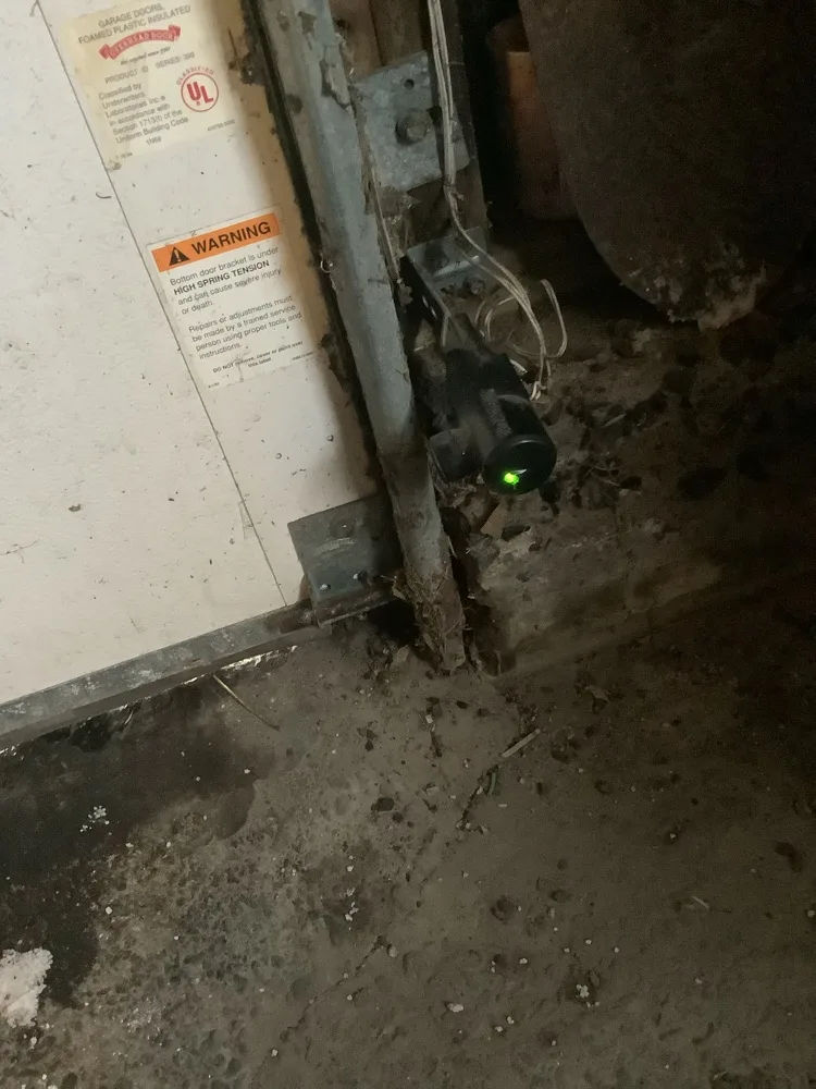 Bottom corner of a garage door at floor level, its bottom bracket carrying a high spring tension warning label beside a photo-eye sensor glowing green on the jamb
