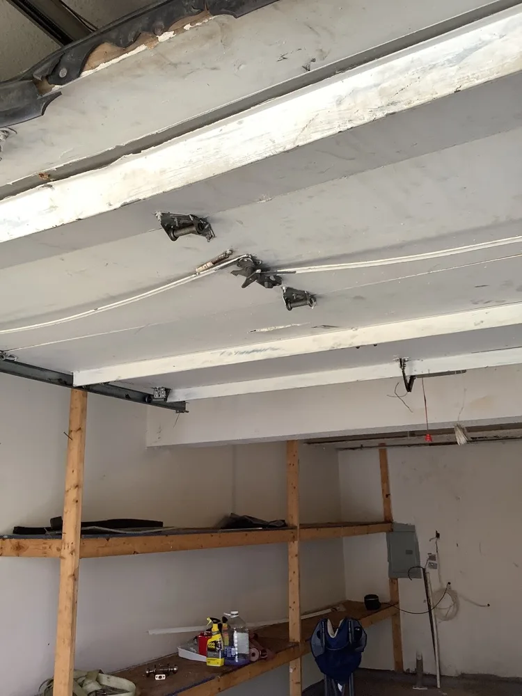 Underside of a garage door parked on its horizontal tracks, hinges and rollers visible along the section seams, above timber storage shelving on the garage wall