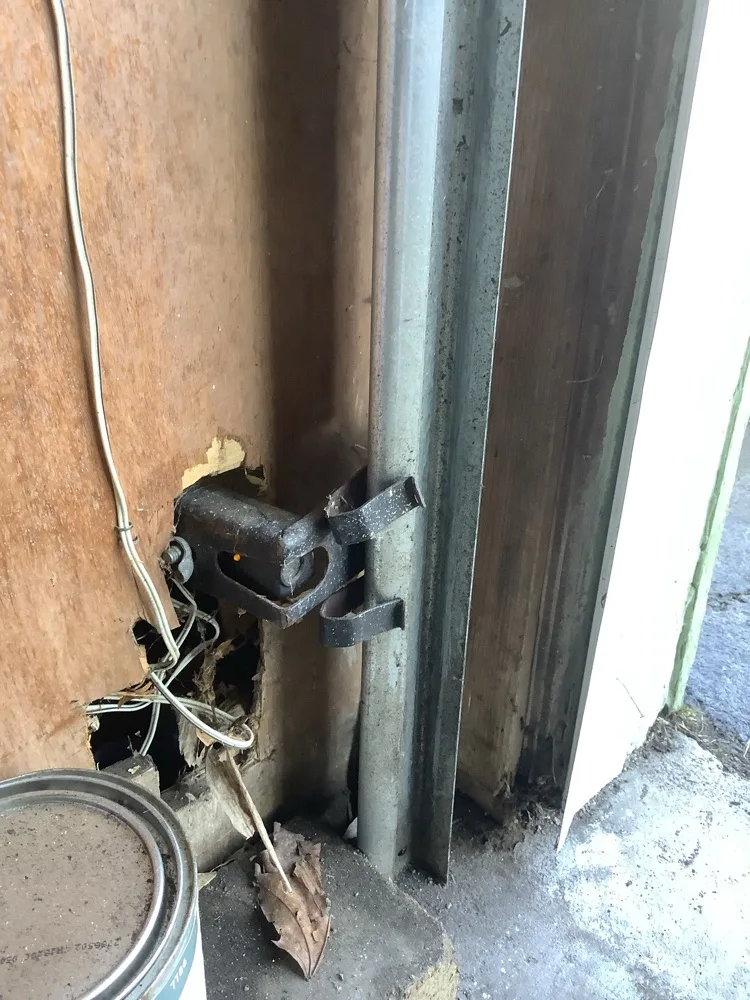 Corroded garage door lock bar and vertical track at a timber jamb, the frame split and rotted around the fixing with sensor wiring pulled loose and a dried leaf on the threshold
