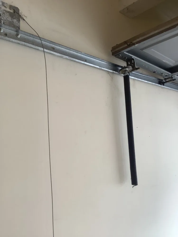 Broken extension spring hanging straight down loose from its track bracket beside a garage door, with the safety cable dangling slack alongside it