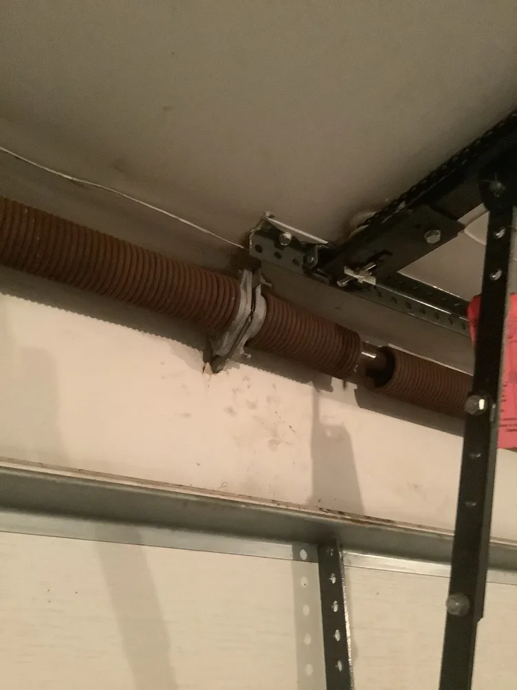 Heavily rusted torsion spring at its centre bracket above a garage door, the coils gone orange with corrosion, alongside the opener rail hanger