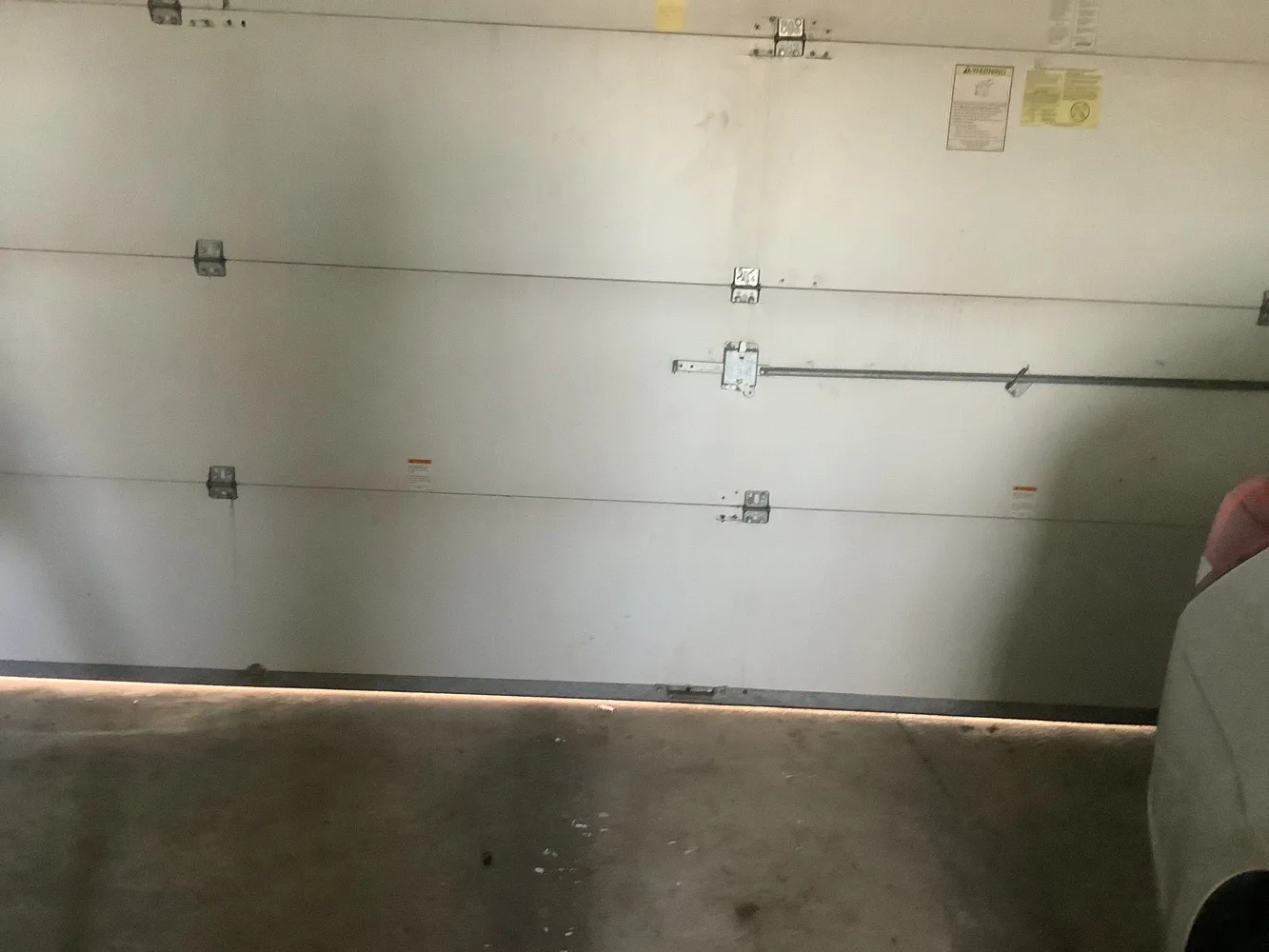 Closed flat white garage door seen from inside a garage, a steel reinforcement strut bolted across it and daylight showing in a line along the bottom seal