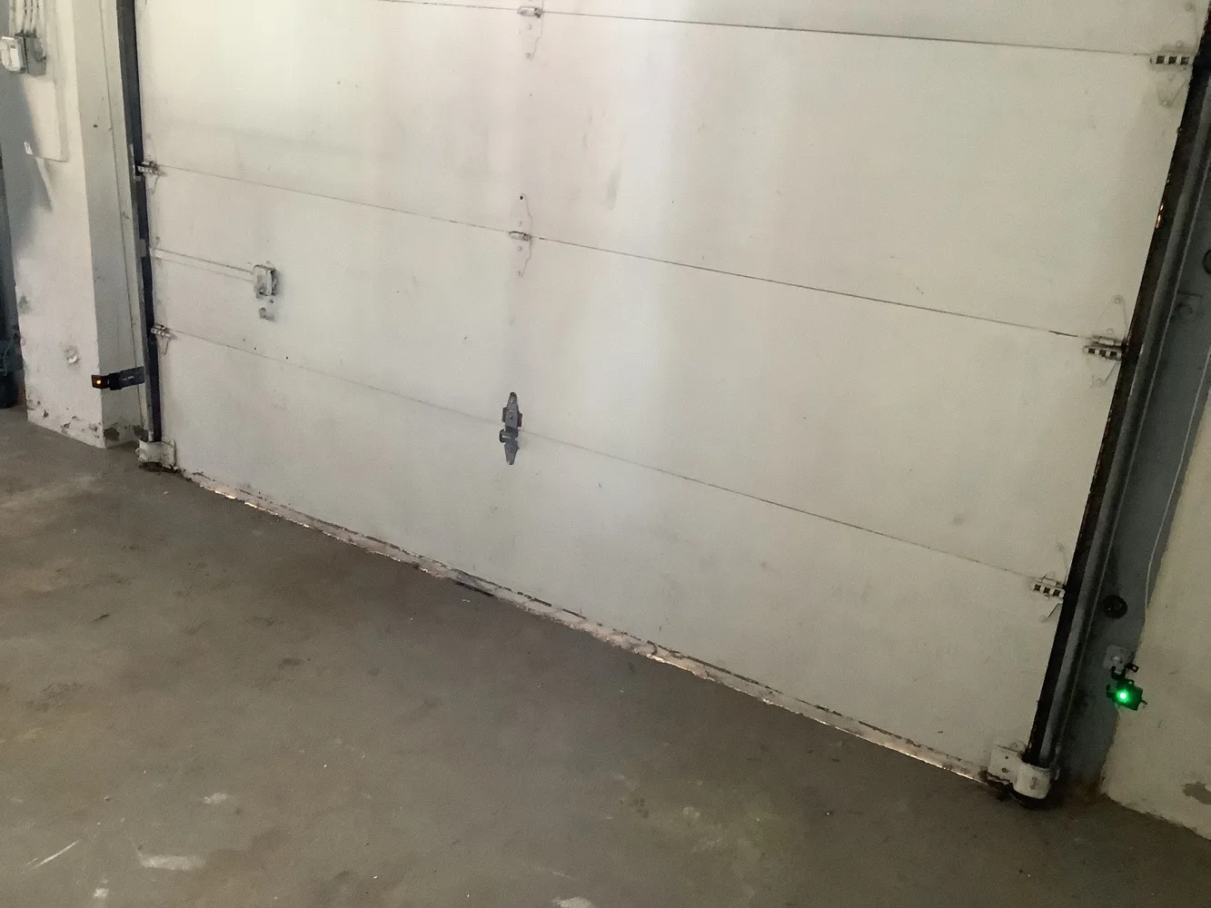 Closed white garage door from inside with a lifting, damaged bottom seal and a photo-eye sensor glowing green low on the right-hand jamb