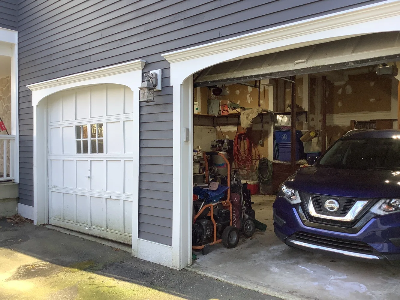 Daylit grey clapboard house with two garage bays, the left door closed with an eight-light window panel and the right bay standing open on a parked car and stored equipment